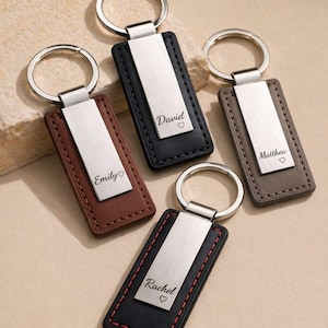 Puede incluir: Varios llaveros personalizados. Cada llavero presenta una base rectangular de cuero en marrón, negro y gris, con una placa de metal plateado. Las placas están grabadas con nombres como "Emily", "David", "Matthew" y "Rachel", cada uno seguido de un símbolo de corazón.