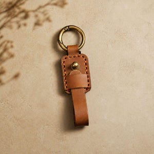 Könnte beinhalten: Ein brauner Leder-Schlüsselanhänger mit einem messingfarbenen Verschluss und Niete. Der Schlüsselanhänger hat ein rechteckiges Lederstück mit genähten Kanten und einer Schlaufe zum Befestigen von Schlüsseln. Der Hintergrund ist ein neutrales Beige.