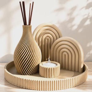 Puede incluir: Un conjunto beige que consta de una bandeja redonda acanalada, un jarrón con textura en espiral con varillas difusoras marrones, dos piezas decorativas en arco con patrones de costillas concéntricas y un pequeño portavelas circular.