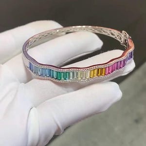 Può includere: Un bracciale rigido incernierato color argento con un design ondulato e smerlato. Il bracciale è incastonato con una fila di pietre preziose taglio baguette in un gradiente arcobaleno, tra cui pietre azzurre, verdi, gialle, arancioni, rosa e viola.
