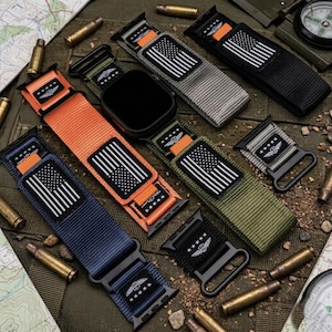 May include: A collection of nylon watch bands in various colors, including orange, navy blue, and black. Each band features a white and black American flag patch and a military-style emblem. The bands are displayed with a black smartwatch and scattered ammunition.
