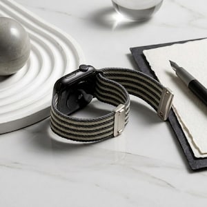 May include: A black smartwatch featuring a woven fabric band with alternating dark gray and light beige horizontal stripes. The watch band includes polished silver-toned metal hardware connectors and a matching silver-toned metal sliding buckle.
