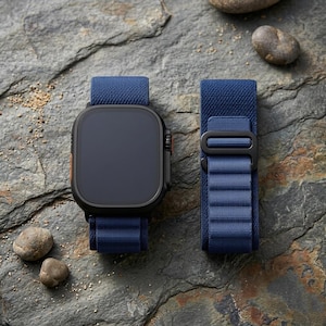 May include: A black smartwatch with a dark blue screen and a matching blue fabric band. The band is detached and lies next to the watch. The watch has an orange accent on the side. The watch and band are on a textured, gray surface.