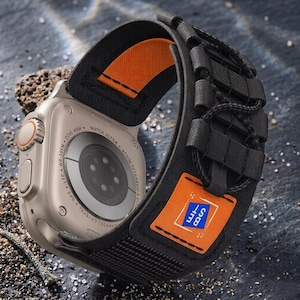 May include: A close-up of a silver smartwatch with a black and orange band. The watch has a digital display and a black back with text. The band has a small orange tag with blue text and a white border. The watch is on a textured surface.