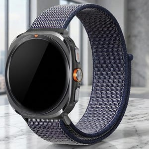 Puede incluir: Un reloj inteligente con caja circular gris oscuro y pantalla negra. El reloj cuenta con una correa de nailon azul oscuro texturizada. En el lateral de la caja, hay un botón circular naranja y dos botones grises adicionales para navegar.