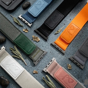 May include: An assortment of watch bands in various colors, including blue, black, orange, green, gray, and pink. Each band features a leather patch with the word "FEARLESS" and a stylized infinity symbol. The bands are displayed on a textured gray surface.