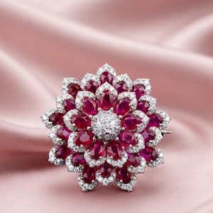 May include: A floral-shaped brooch featuring multiple layers of oval-cut deep red rubies. Each ruby is surrounded by a border of small, clear, sparkling white stones. The center of the brooch is a cluster of small, round, clear white stones.