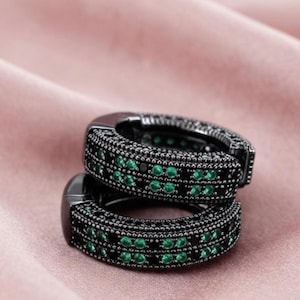 May include: A pair of black hoop earrings featuring a thick band design. The surface of each earring is encrusted with two parallel rows of small, vibrant green circular stones, interspersed with tiny black stones in a textured, pave-style setting.