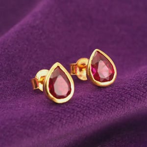 May include: A pair of stud earrings featuring teardrop-shaped red gemstones set in polished yellow gold bezels. The earrings have a classic post-back design, showcasing the vibrant crimson colour of the stones and the warm metallic lustre of the gold settings.