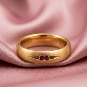 Könnte beinhalten: Ein goldener Ring mit gebürstetem, mattem Finish, der zwei kleine, runde, leuchtend rote Rubine zeigt, die horizontal in der Mitte gefasst sind. Die Steine befinden sich in einer schmalen, spitz zulaufenden horizontalen Kerbe.