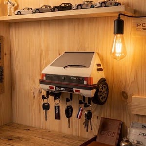 Puede incluir: Un llavero Peugeot 205 GTI blanco y naranja montado en una pared de madera, con llaves colgando debajo. El llavero tiene una rueda de coche en el lateral. Modelos de coches en miniatura sobre una estantería. Una bombilla cuelga del techo.
