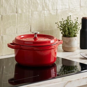 May include: A vibrant red cast iron casserole dish with a lid and handles is on a black hob. A small potted herb, a black bottle, and a wooden pepper mill are in the background. A stainless steel spoon is on the counter.