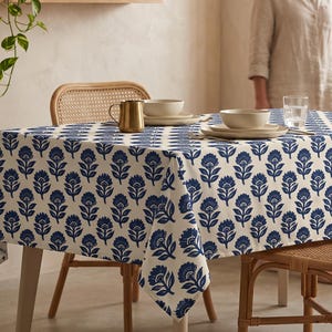 May include: A rectangular tablecloth featuring a repeating dark blue floral pattern on an off-white background. The fabric drapes over a dining table, which is set with a gold-toned metal pitcher, stacked ceramic bowls, and a clear glass.