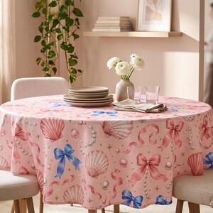 May include: A round tablecloth featuring a light pink background with a repeating pattern of pink and blue bows, white seashells, and strings of pearls. The fabric drapes over a circular table, which is set with a stack of plates and a vase.