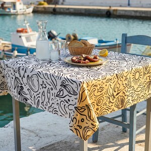 May include: A square table covered with a white and yellow tablecloth featuring a black line-art pattern of suns, fish, shells, and olive branches. On the table sits a glass carafe, a small glass, a bread basket, and a plate with cooked octopus.