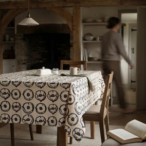 May include: A square tablecloth featuring a repeating pattern of stylized dark blue and brown poppy flowers on a cream-colored fabric. The tablecloth is draped over a wooden table, which also holds a white butter dish and a ceramic mug.