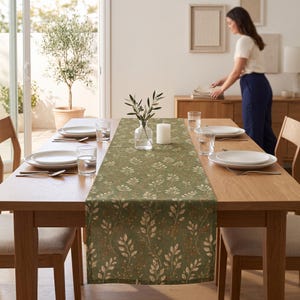 May include: A rectangular table runner featuring a sage green background with a repeating pattern of white leaves and small brown berries, centered on a wooden dining table set with white plates, clear glasses, and a white pillar candle.