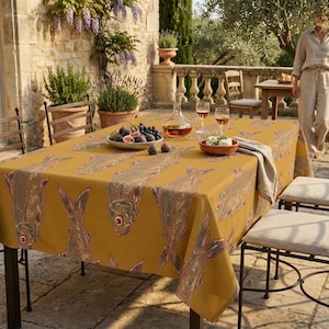 May include: A mustard yellow tablecloth featuring a repeating pattern of stylized fish in shades of brown, blue, and green. The table is set with a glass carafe, two wine glasses, a bowl of fresh figs and grapes, and a small bowl of cheese.