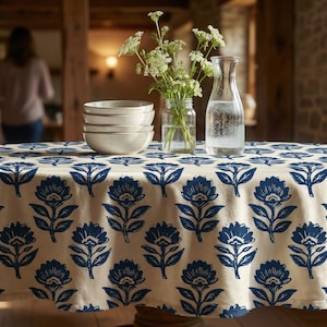 May include: A round tablecloth with a cream background and a repeating pattern of blue floral designs. The table is set with white bowls, a glass water pitcher, and a small vase of flowers.