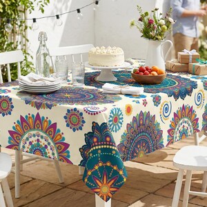 May include: A table set for an outdoor gathering, featuring a cream-colored tablecloth with a vibrant, teal, purple, and yellow mandala pattern. White chairs surround the table, which is set with plates, glasses, and a cake. A vase of flowers and a bowl of fruit add to the festive atmosphere.