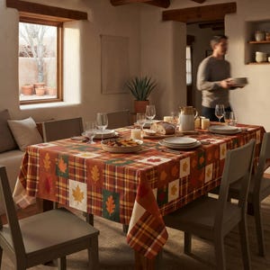 May include: A rectangular tablecloth featuring a patchwork design of autumn-themed plaid patterns and various colorful leaf motifs in shades of orange, yellow, red, and green, draped over a dining table set with plates, glasses, and a pitcher.