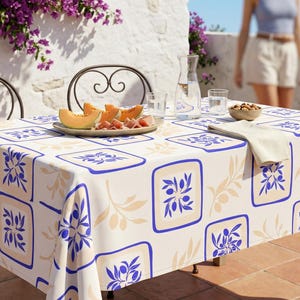 May include: A rectangular tablecloth with a repeating pattern of blue squares with olive branch designs on a cream background. The table is set with a plate of melon and prosciutto, glasses, and a bowl of nuts. The table is set outdoors.