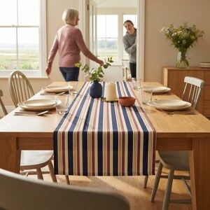 May include: A rectangular table runner featuring vertical stripes in navy blue, burgundy, and cream. The runner is centered on a light oak dining table set with white plates, clear glasses, a dark blue vase with greenery, and a pillar candle.
