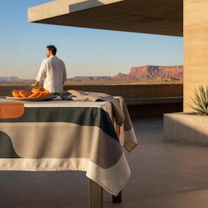 May include: A modern outdoor dining scene featuring a table with a patterned tablecloth in shades of orange, green, and gray. A glass and a plate of sliced oranges sit on the table. A person stands in the background, overlooking a desert landscape.