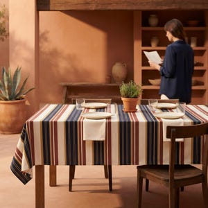 May include: A rectangular tablecloth featuring vertical stripes in navy blue, terracotta red, mustard yellow, and cream. The table is set with four white ceramic plates, four clear glass tumblers, and a potted green herb in a terracotta pot.