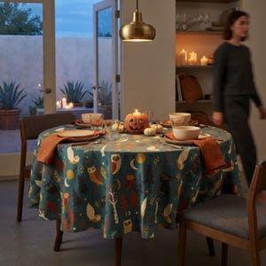 May include: A round dining table covered with a teal tablecloth featuring a pattern of owls, moons, and trees. The table is set with plates, bowls, and orange napkins. A carved jack-o'-lantern sits in the center, surrounded by small white pumpkins.