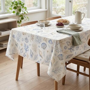 May include: A rectangular tablecloth featuring a repeating pattern of blue and beige seashells, starfish, and coral branches on a white background. The fabric drapes over a wooden table set with ceramic bowls, a pitcher, and a folded green napkin.