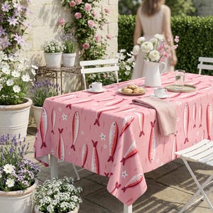 May include: A rectangular tablecloth in a soft pink hue featuring a repeating pattern of stylized red and white fish and small white flowers. The fabric drapes over a table set with a white pitcher of flowers, plates, and cups.