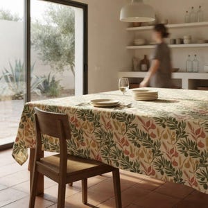 May include: A rectangular tablecloth featuring a botanical pattern of green olive branches, wheat stalks, and small reddish-brown figs on a cream-colored background. The fabric drapes over a wooden table, accompanied by a wooden chair and a wine glass.