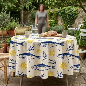 May include: A round tablecloth featuring a repeating pattern of blue fish, yellow lemon slices, and small blue sprigs on a cream-colored background. The tablecloth is draped over a table set with a glass pitcher, two mugs, and a wooden cutting board.