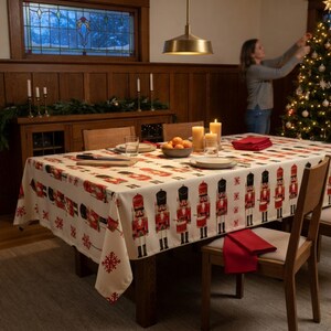 May include: A rectangular tablecloth featuring a repeating pattern of red and white nutcracker figures and red snowflakes on a cream background. The fabric covers a wooden dining table set with plates, a bowl of oranges, and candles.