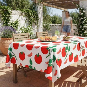 May include: A white tablecloth with a repeating pattern of red tomatoes and green leaves covers a wooden table. The table is set outdoors with food and drinks, creating a summery dining scene.