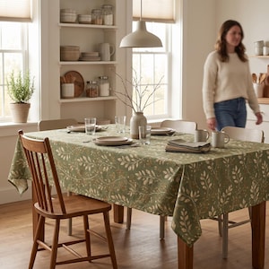 May include: A rectangular dining table covered with a sage green tablecloth featuring a repeating pattern of cream-colored leaves and small brown berries. The table is set with ceramic plates, clear drinking glasses, and a central vase with branches.