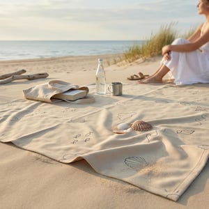 May include: A beige beach towel featuring a black line-art pattern of seashells, waves, and small fish. The rectangular towel is spread out on sand and includes a matching beige tote bag, a clear glass water bottle, and a small metal cup.