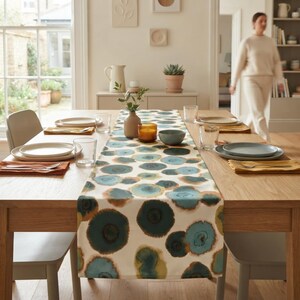 May include: A rectangular table runner featuring a repeating pattern of abstract circular shapes in shades of teal, blue, and mustard yellow on a cream background, laid across a light wood dining table set with white plates and clear glassware.