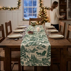 May include: A long rectangular table runner featuring a green and cream toile-style print of a winter forest landscape with deer, a horse-drawn sleigh, and pine trees, centered on a wooden dining table with white plates and glasses.