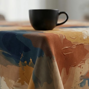 May include: A black ceramic coffee mug sits on a tablecloth with an abstract design. The tablecloth features brushstrokes of blue, orange, and yellow on a cream background. The scene is set indoors with soft lighting.
