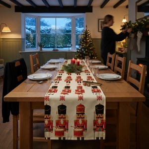 May include: A cream-colored table runner featuring a repeating pattern of red and black nutcracker figures, each holding a drum or spear, interspersed with small red snowflake motifs, laid flat across a wooden dining table.