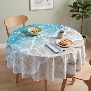 May include: A round tablecloth featuring a high-resolution print of a sandy beach with turquoise ocean waves washing onto the shore. The design includes realistic details of sea foam, wet sand, and a large seashell. A bowl of fruit, a mug, and a pastry sit on top.