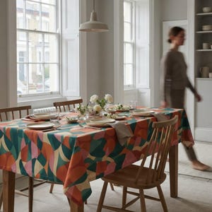 May include: A rectangular dining table covered with a tablecloth featuring a bold, multicolored geometric pattern in shades of orange, teal, pink, and cream. The table is set with white plates, clear glassware, and small vases with white flowers.