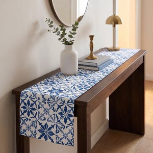 May include: A rectangular table runner featuring a repeating blue and white geometric star pattern. The fabric runner is draped over a dark wood console table, showcasing a crisp, high-contrast design with intricate floral-inspired motifs.