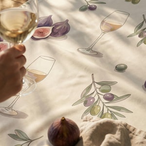 May include: A cream-colored tablecloth featuring a repeating pattern of purple figs, green olive branches, and glasses of white wine. The fabric displays a detailed illustration of whole and halved figs alongside delicate wine glasses and olives.