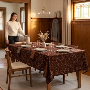 May include: A rectangular dining table covered with a dark brown tablecloth featuring an orange and brown leaf pattern. The table is set with white plates, clear drinking glasses, brown cloth napkins, and a central vase filled with dried wheat stalks.