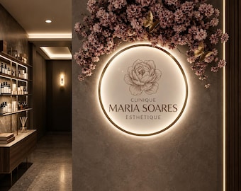 以下が含まれることがあります： ピオニーの線画イラストと「CLINIQUE MARIA SOARES ESTHÉTIQUE」の文字が描かれた、円形のバックライト付きウォールサイン。ピンクの花の枝と2つの金色の蝶のアクセントで飾られた暗い壁に取り付けられています。