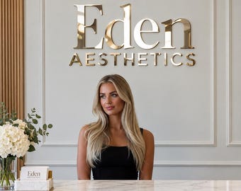 以下が含まれることがあります： 壁に金色の金属文字でEden AESTHETICSと書かれた受付エリア。手前には白い大理石のカウンターがあり、白いアジサイが入ったガラスの花瓶と、Eden AESTHETICSと記された小さな白いカードホルダーが置かれています。