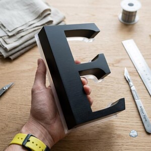 Op de afbeelding: Een driedimensionale letter E met een matzwarte voorzijde en een transparante acryl achterzijde. De letter heeft een dik blokletterontwerp met scherpe randen en een gladde afwerking, vastgehouden in een hand boven een houten werkoppervlak.